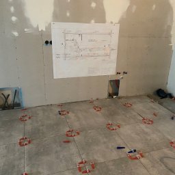 KATRUK Technologies - Szare płytki podłogowe ułożone z użyciem czerwonych i niebieskich klinów poziomujących, ściany wykończone płytami gipsowo-kartonowymi, na ścianie wisi plan instalacji.