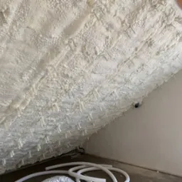 Izolacja poddasza pianką PUR z widocznymi plastikowymi kołkami mocującymi, białe rury wentylacyjne na podłodze, surowa ściana z instalacją elektryczną.