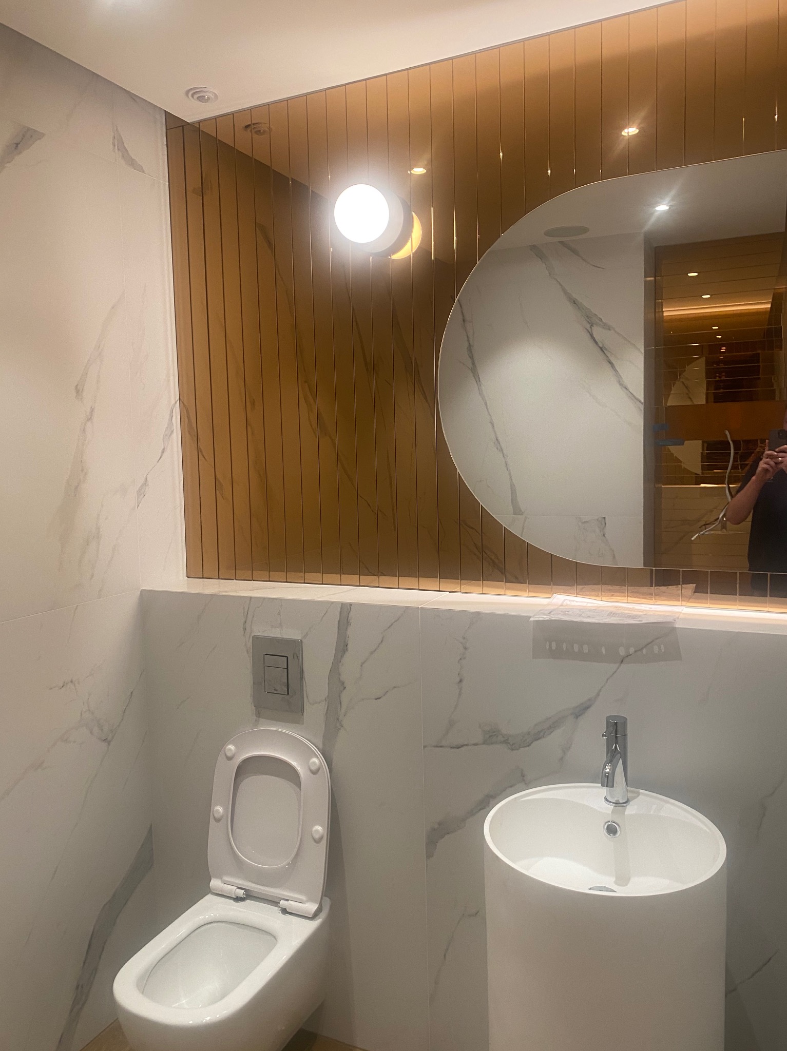 Nowoczesna łazienka we Wrocławiu z marmurowymi płytkami, okrągłym lustrem z oświetleniem LED, wiszącą toaletą z podtynkowym systemem spłukiwania i umywalką na postumencie.