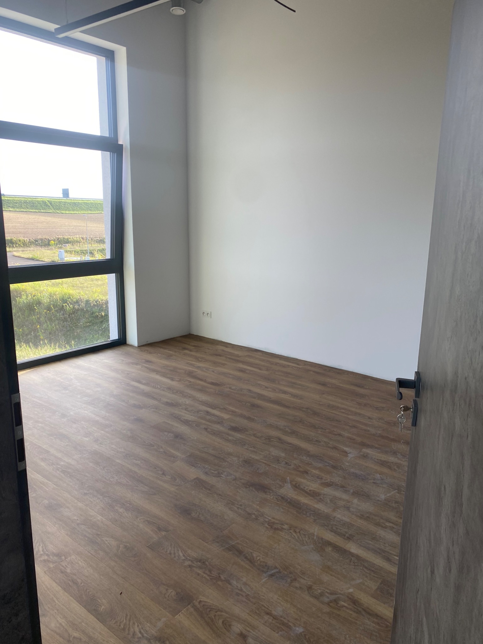 Pusty pokój z panelami podłogowymi w odcieniach brązu, świeżo pomalowanymi białymi ścianami i dużym oknem z widokiem na zewnątrz, klucze wiszące w zamku drzwi.