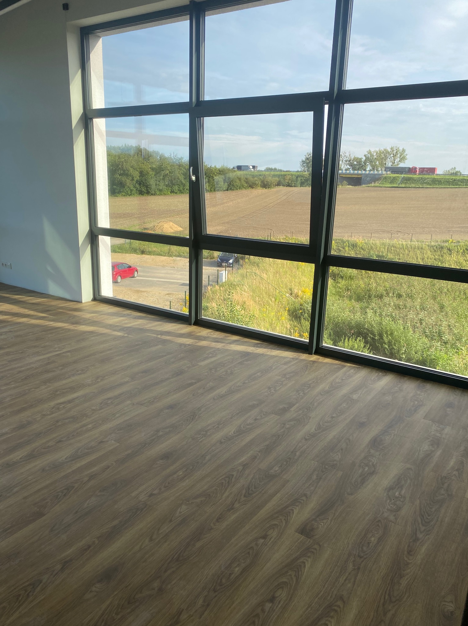 Nowa podłoga z paneli w pustym pokoju z dużym oknem z widokiem na krajobraz wiejski z przejeżdżającymi samochodami.
