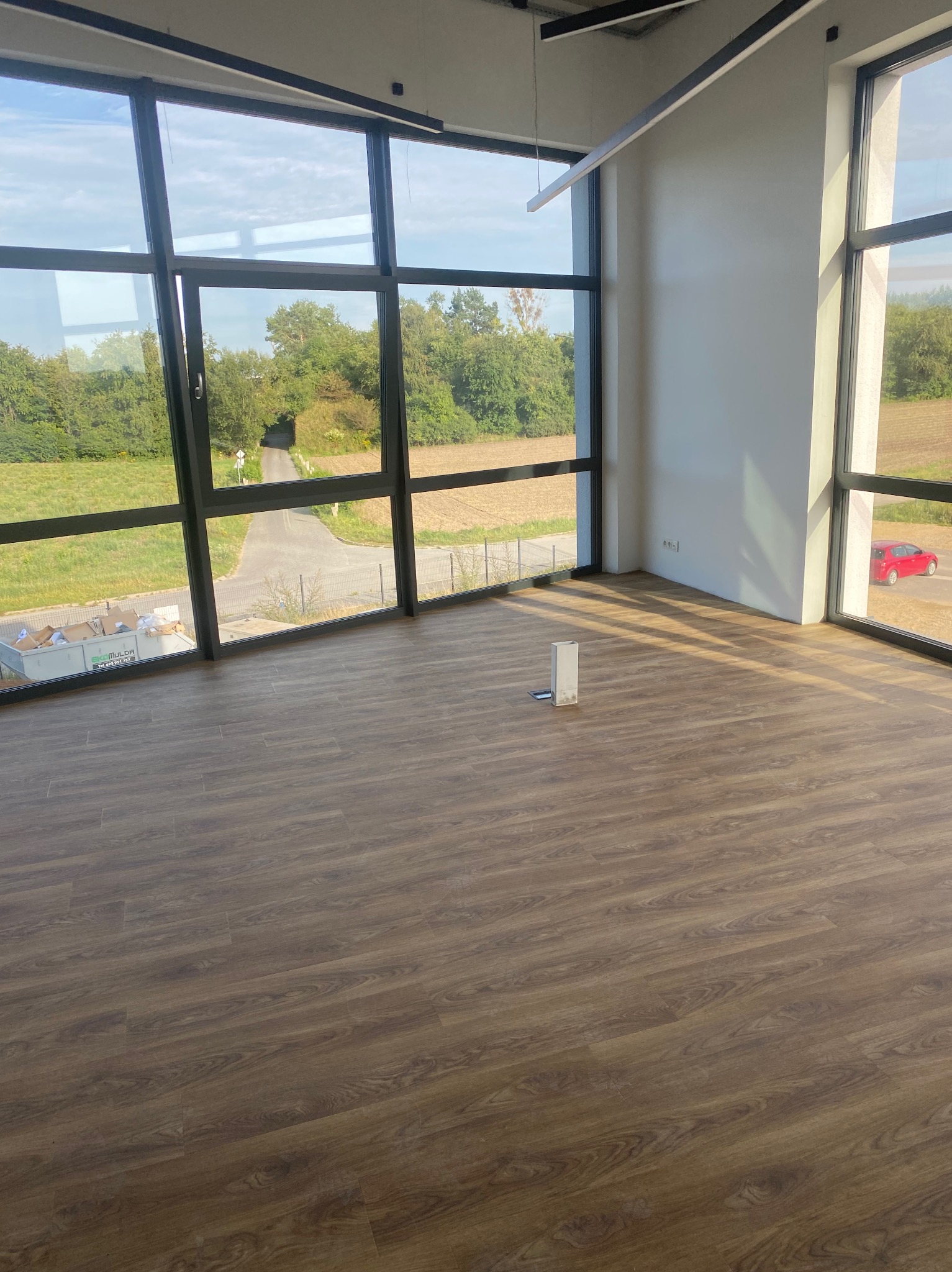 Nowa, pusta przestrzeń biurowa z panelami podłogowymi w odcieniach brązu, widok na zieleń przez duże okna, minimalistyczne oświetlenie sufitowe i białe ściany.