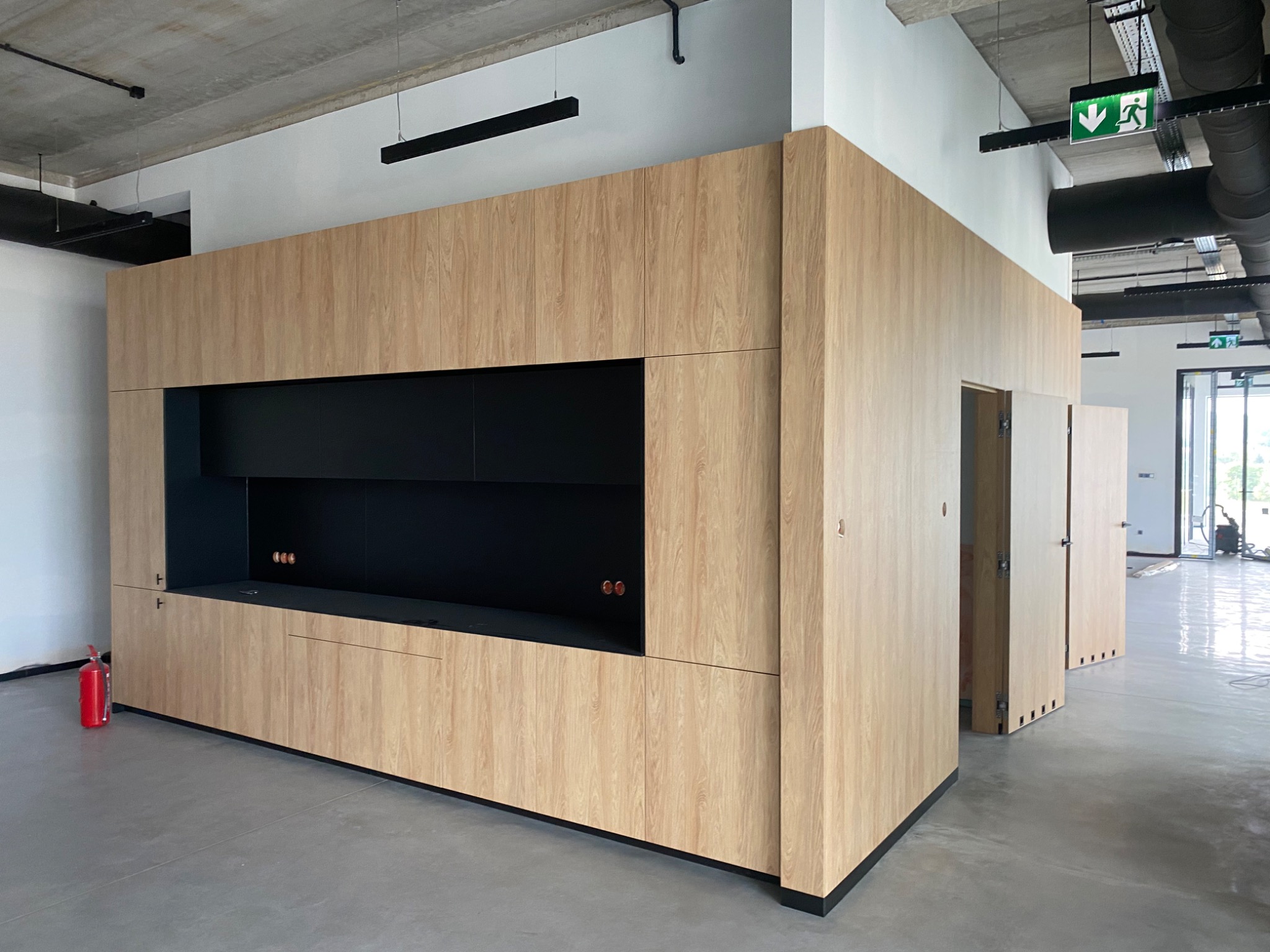 Wykończona zabudowa meblowa z jasnego drewna w biurze, z czarnymi wnękami i gniazdkami, widoczny betonowy sufit i instalacje.