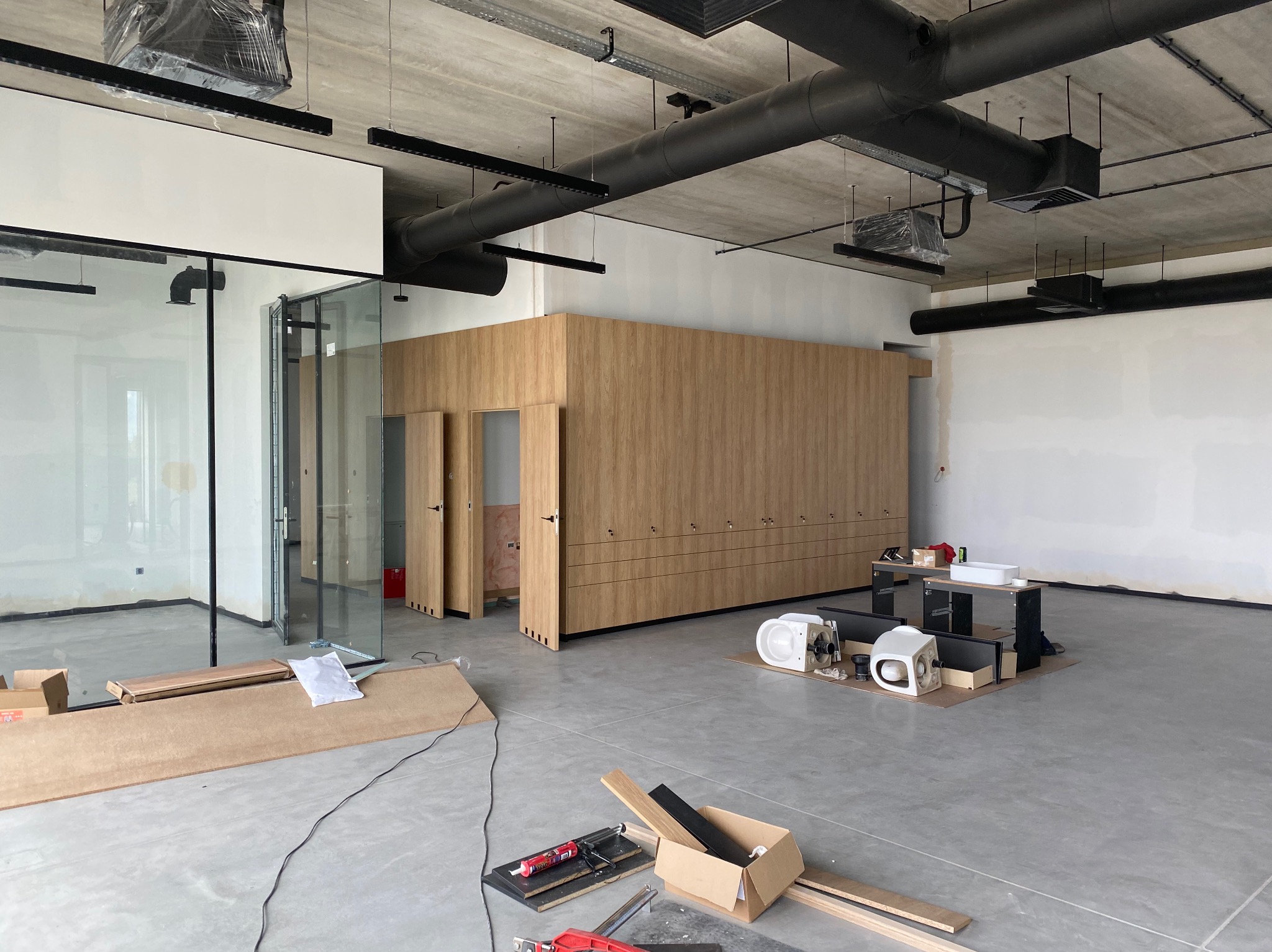 Wnętrze biurowe w trakcie wykańczania: widoczne przeszklone ścianki działowe, zabudowa meblowa z drewna, surowy betonowy sufit z instalacjami oraz narzędzia i materiały budowlane na posadzce.