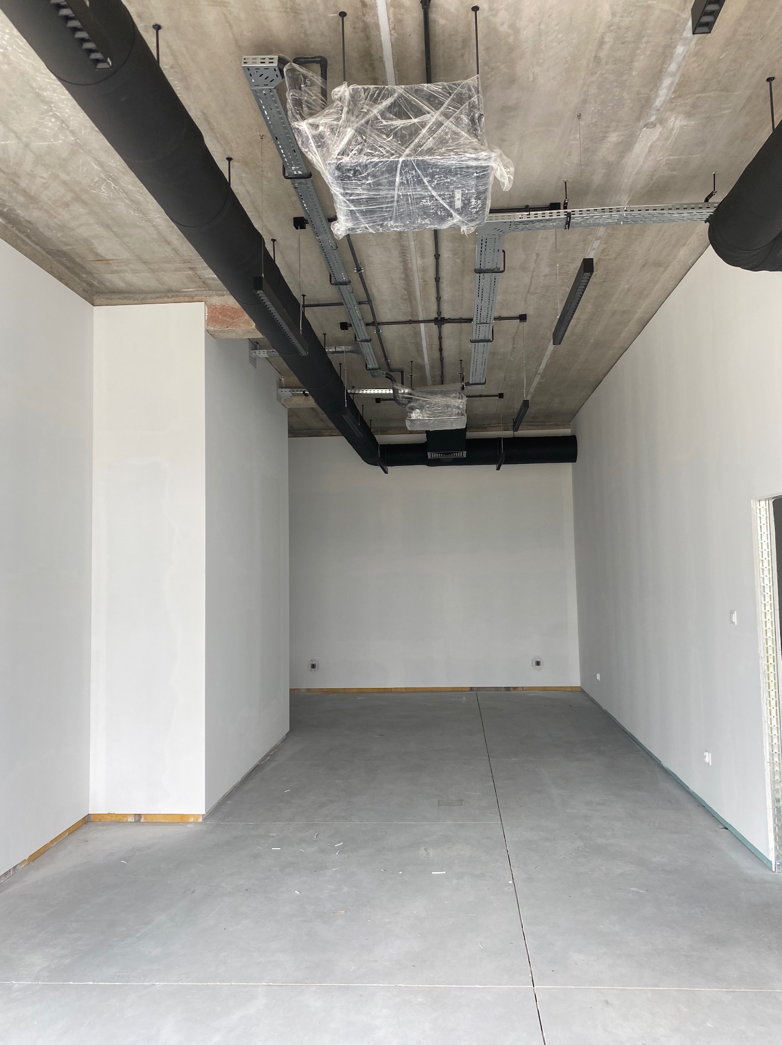 Surowe wnętrze w trakcie prac wykończeniowych, z widocznymi instalacjami pod sufitem, świeżo pomalowanymi ścianami i surową posadzką.