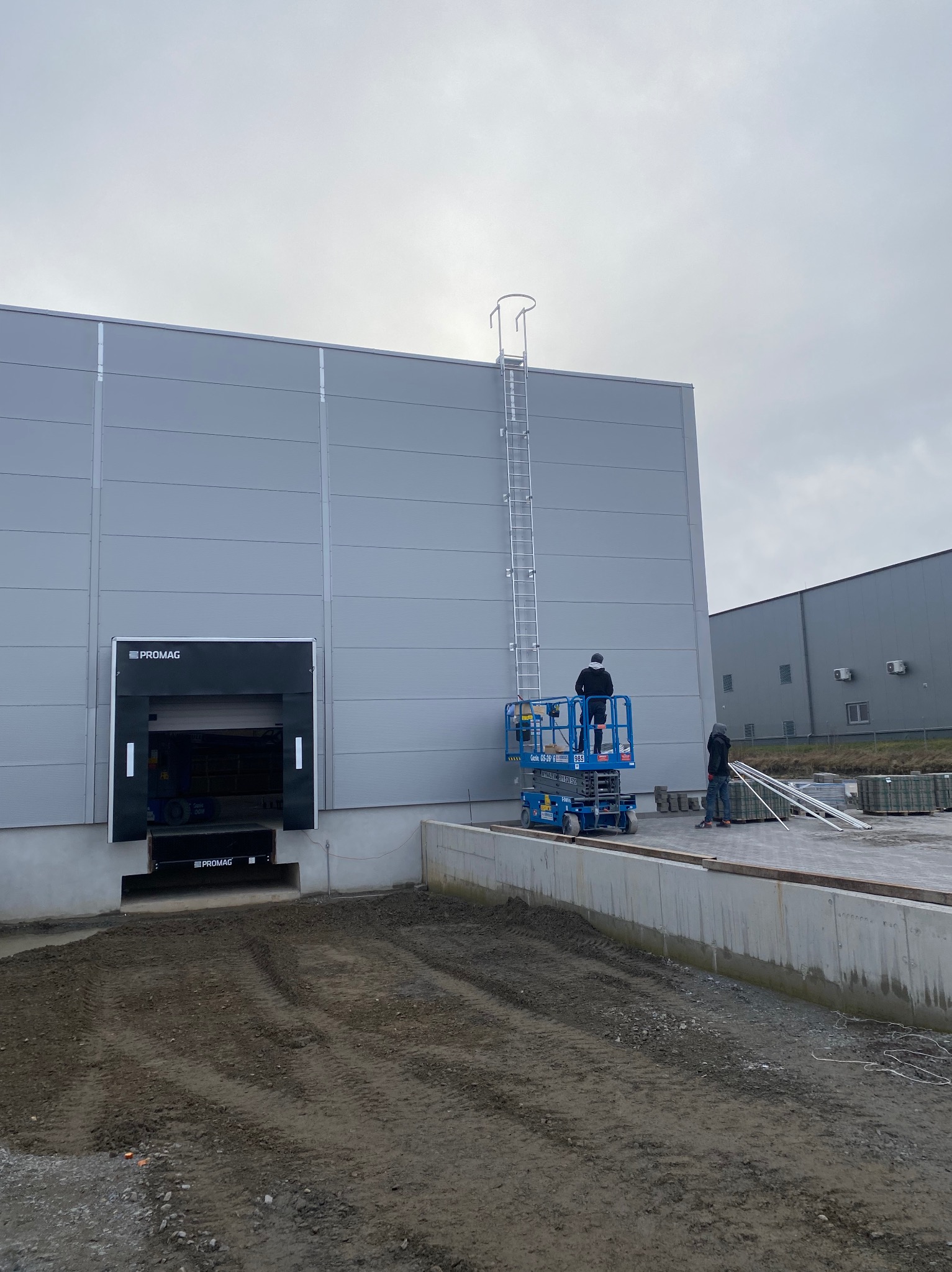 Montaż drabiny pionowej na elewacji hali magazynowej z wykorzystaniem podnośnika koszowego, widoczny pracownik w czarnej kurtce, drugi pracownik na ziemi, elementy elewacji w kolorze szarym.