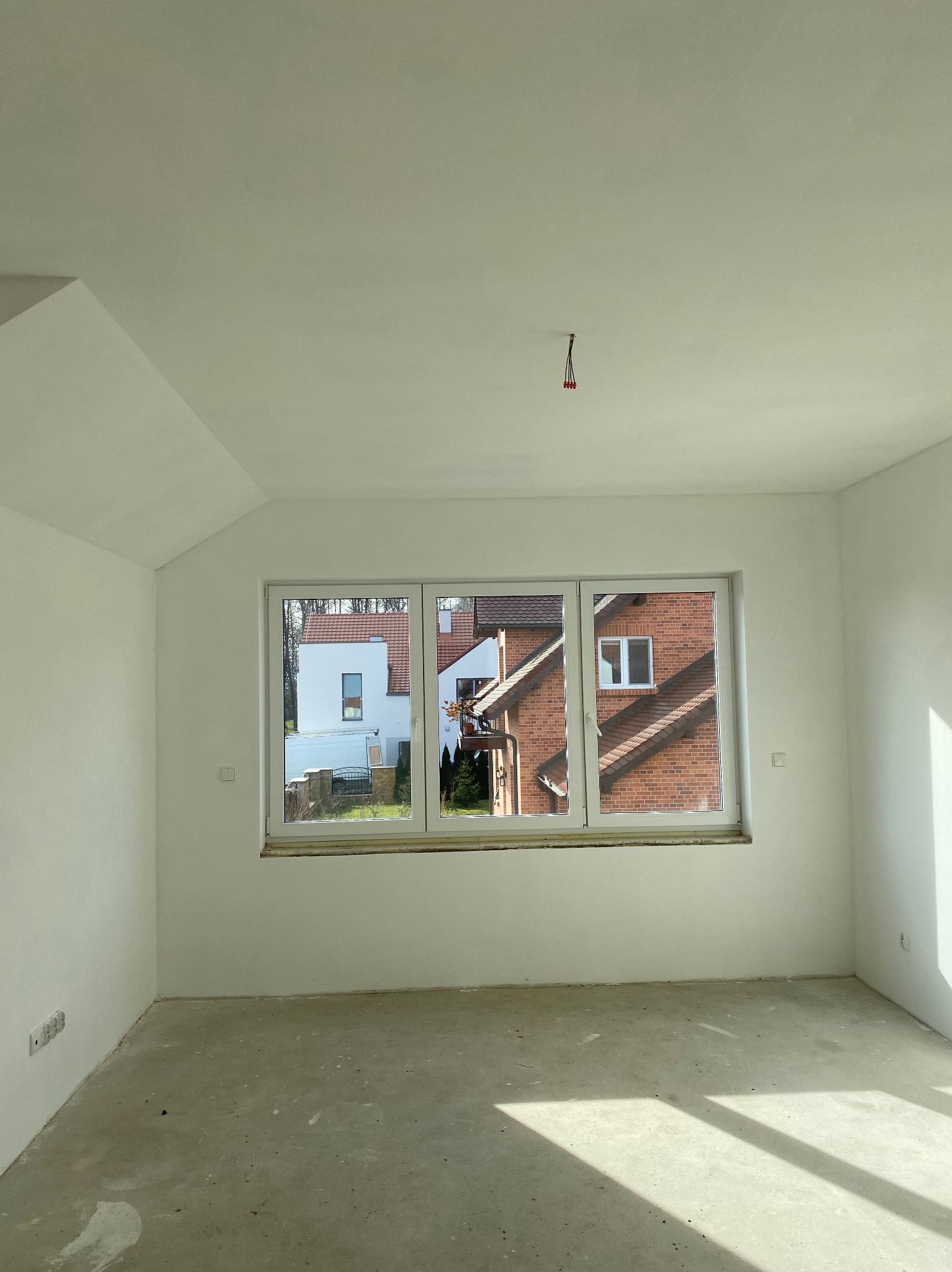 Pusty, pomalowany na biało pokój z surową podłogą, okno z widokiem na domy z czerwonej cegły i białe budynki, w rogu ściany widoczne gniazdka elektryczne.