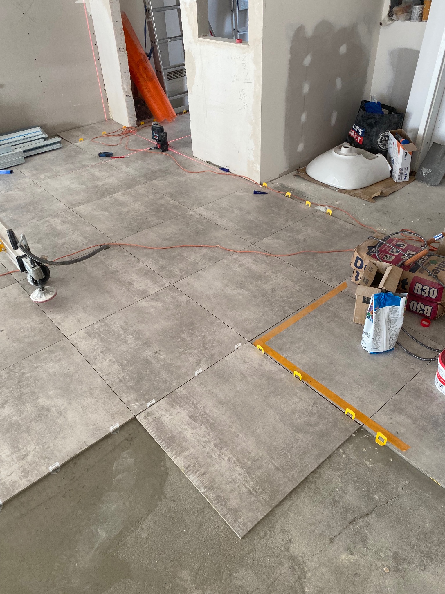 Układanie szarych płytek podłogowych w nowym pomieszczeniu z użyciem poziomicy laserowej i systemu poziomowania płytek, widoczne worki z zaprawą i niezamontowana umywalka.