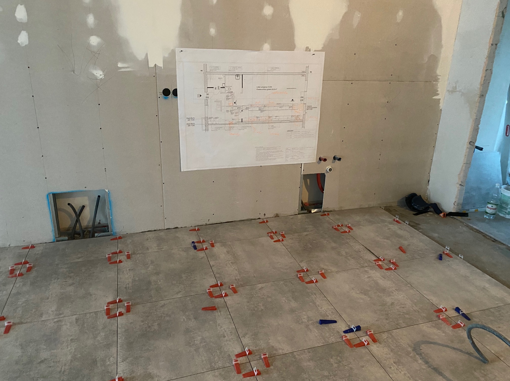 Szare płytki podłogowe ułożone z użyciem czerwonych i niebieskich klinów poziomujących, ściany wykończone płytami gipsowo-kartonowymi, na ścianie wisi plan instalacji.