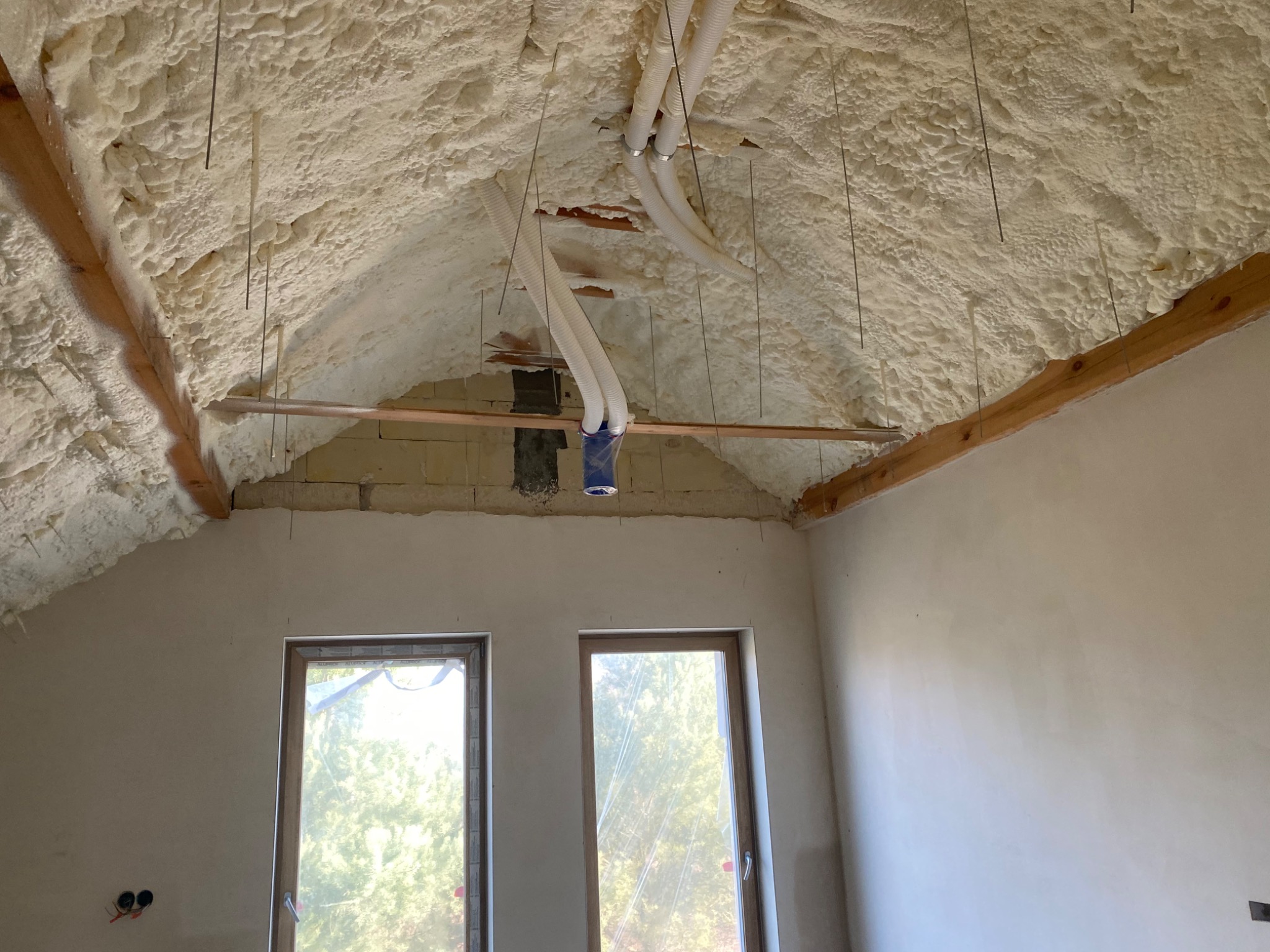 Poddasze w trakcie izolacji pianą poliuretanową, widoczne drewniane elementy konstrukcyjne, wystające przewody wentylacyjne i okna zabezpieczone folią.