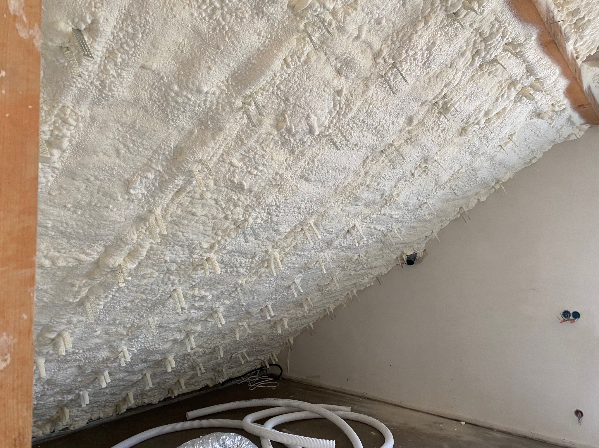 Izolacja poddasza pianką PUR z widocznymi plastikowymi kołkami mocującymi, białe rury wentylacyjne na podłodze, surowa ściana z instalacją elektryczną.