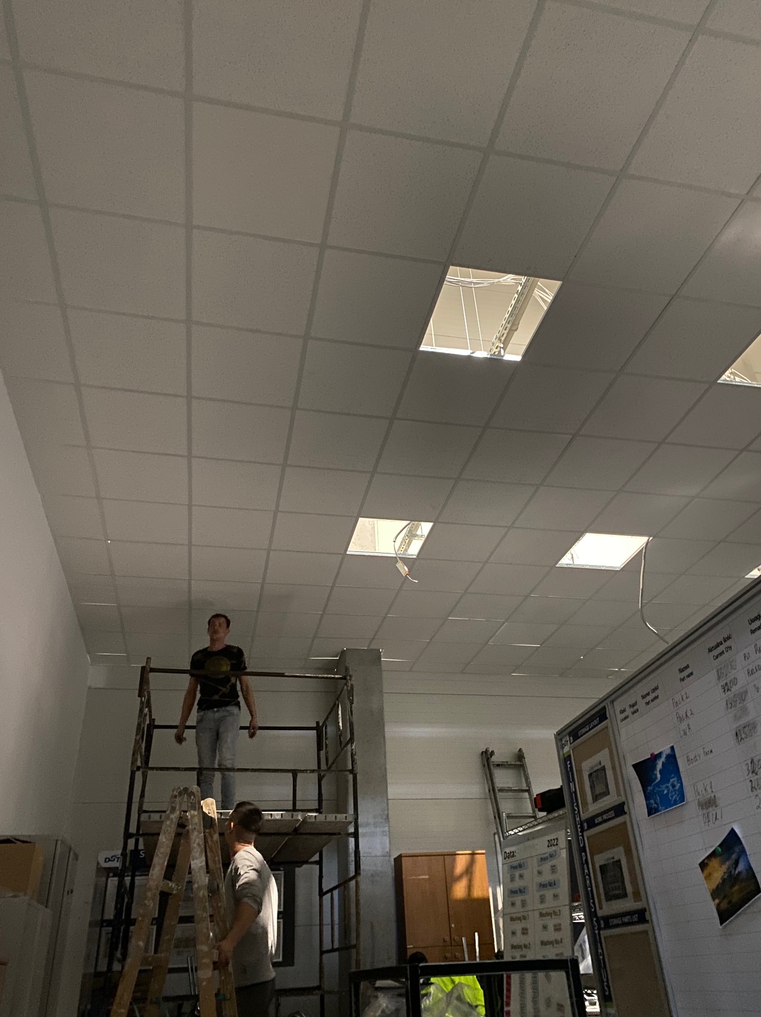 Dwóch mężczyzn na rusztowaniu i drabinie podczas montażu sufitu podwieszanego z widocznymi otworami na oświetlenie.