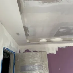 Nierównomiernie nałożona szara gładź na suficie z obniżeniem, widoczne ślady szpachlowania na ścianie, zabezpieczone folią drzwi w tle, fioletowa ściana w dolnej części kadru.