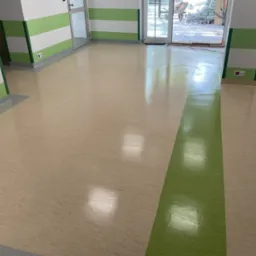 Wypolerowana podłoga z wykładziny PCV w odcieniach beżu i zieleni, świeżo po ułożeniu w pomieszczeniu z pomalowanymi na biało-zielono ścianami i widocznym grzejnikiem oraz otwartymi drzwiami...