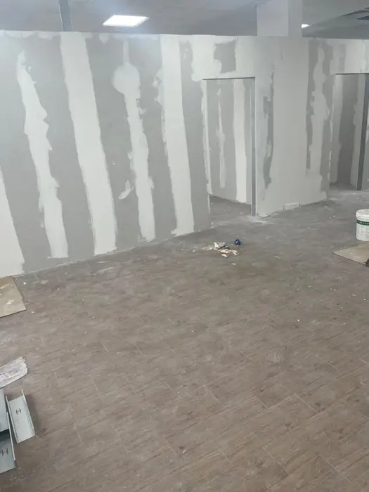 Wnętrze w trakcie remontu: ściany z nałożonymi pasami szpachli, przygotowane do dalszej obróbki, podłoga wyłożona płytkami imitującymi drewno, widoczne profile metalowe i resztki materiałów...