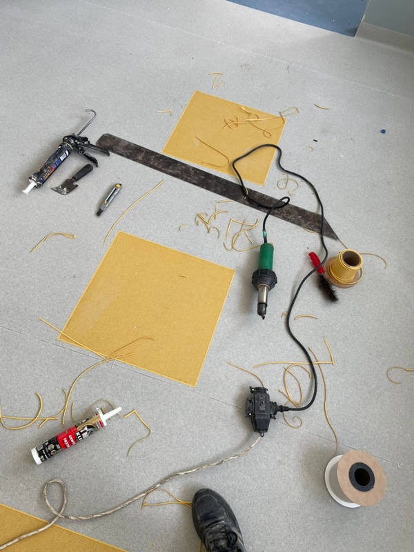 Fragment podłogi z żółtymi kwadratami wykładziny, narzędzia do spawania wykładzin PVC, pistolet do kleju, skrawki spoiwa i stopa w czarnym bucie w rogu kadru.