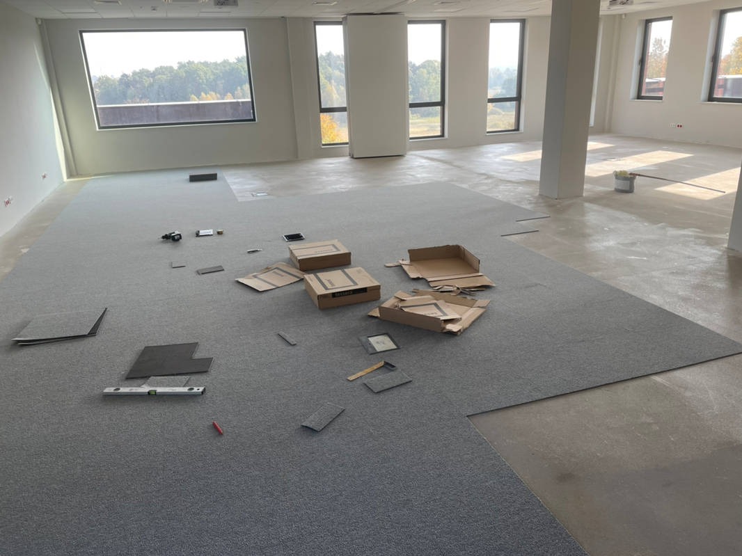 Szare płytki wykładzinowe ułożone częściowo na podłodze w jasnym biurze, widoczne narzędzia, poziomica, kartony po płytkach i niezabudowana część podłogi