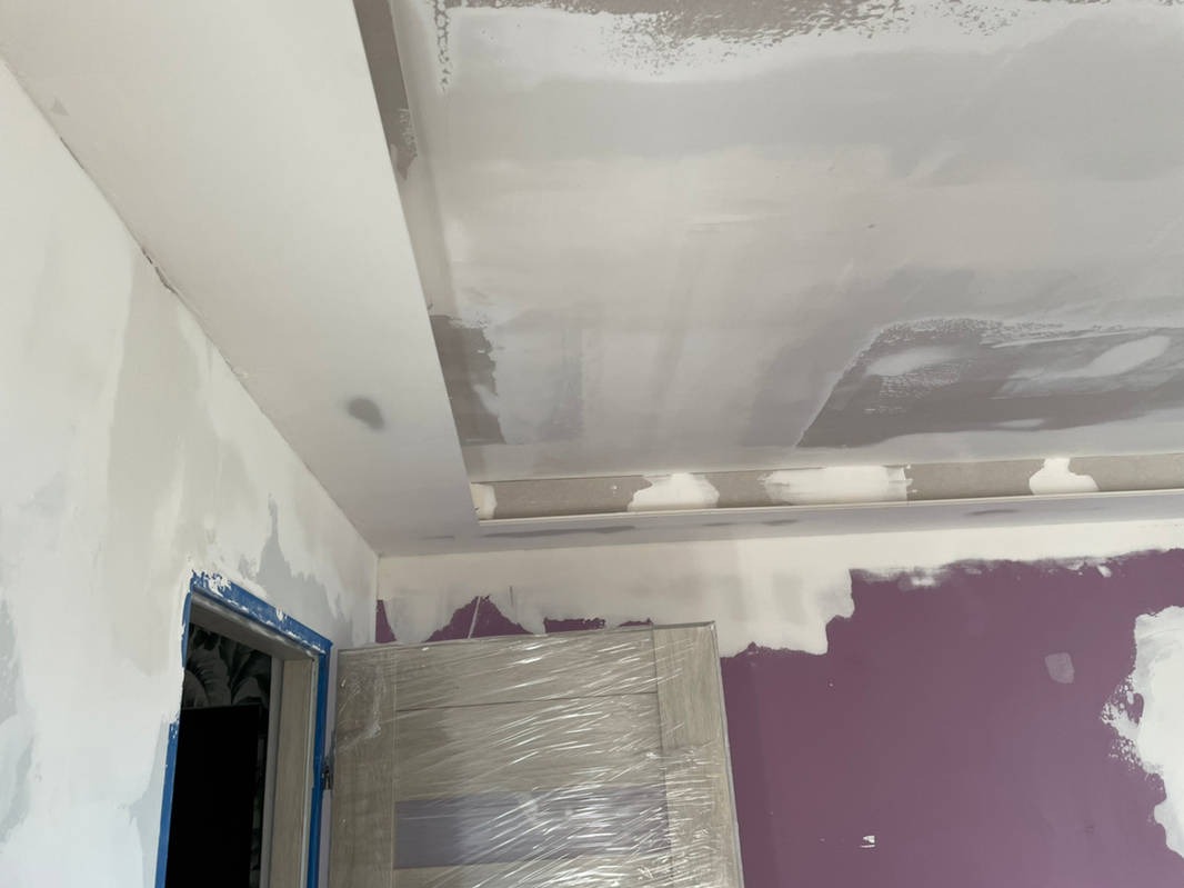 Nierównomiernie nałożona szara gładź na suficie z obniżeniem, widoczne ślady szpachlowania na ścianie, zabezpieczone folią drzwi w tle, fioletowa ściana w dolnej części kadru.