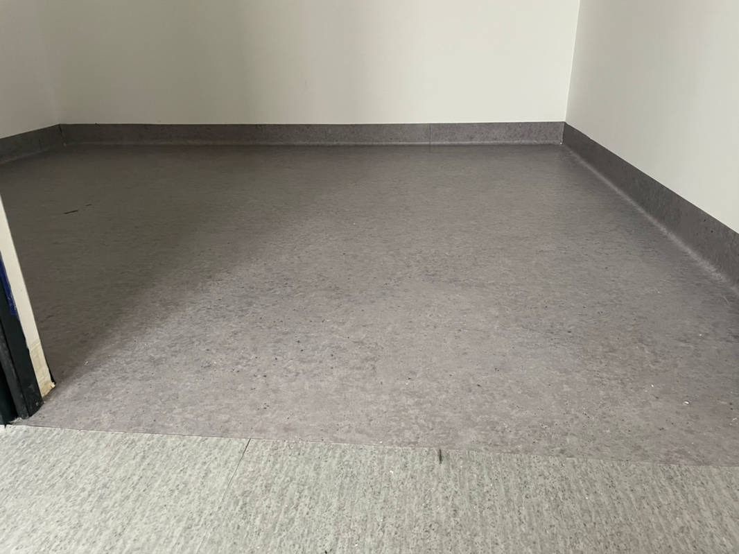 Szara, jednolita wykładzina PCV z listwami przypodłogowymi w rogu pomieszczenia w Feliksowie, widoczny próg drzwiowy.