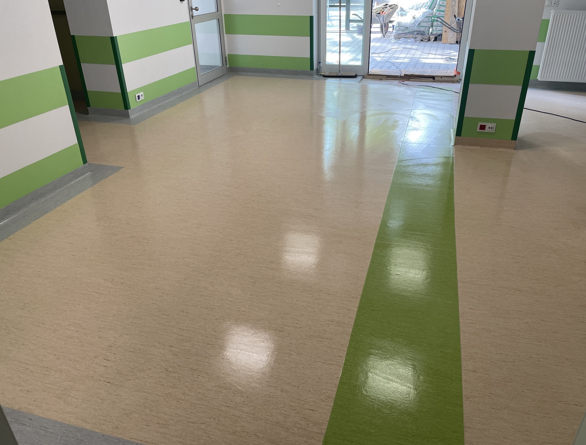 Wypolerowana podłoga z wykładziny PCV w odcieniach beżu i zieleni, świeżo po ułożeniu w pomieszczeniu z pomalowanymi na biało-zielono ścianami i widocznym grzejnikiem oraz otwartymi drzwiami...