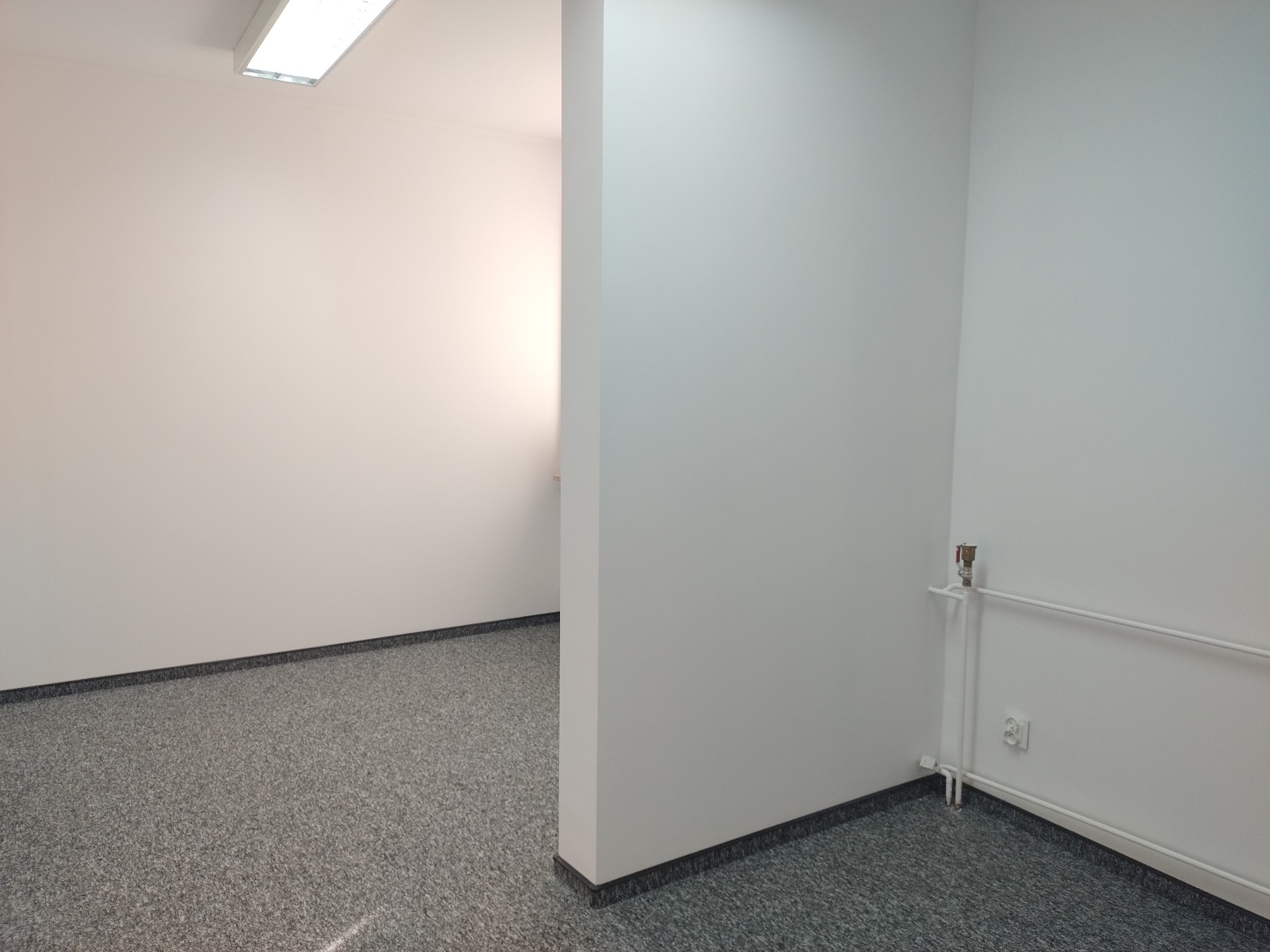 Puste, świeżo odmalowane pomieszczenie biurowe z szarą wykładziną dywanową i białymi ścianami, widoczny grzejnik przy ścianie.