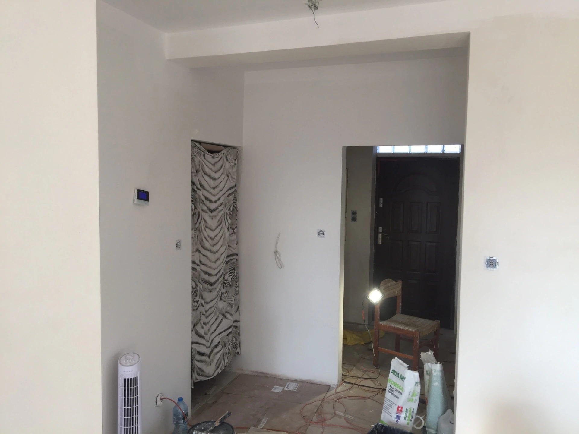 Wnętrze pokoju w trakcie odświeżania, ściany pomalowane na biało, widoczne kable elektryczne wystające ze ściany, podłoga częściowo zabezpieczona kartonem, w tle krzesło z lampą oświetlającą...