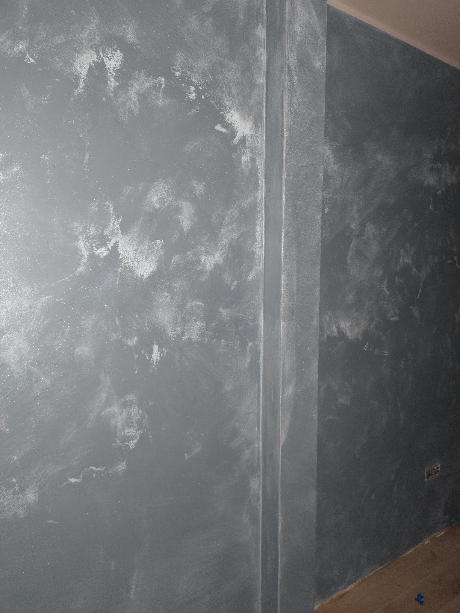 Ściana po nałożeniu szarej farby z efektem dekoracyjnym, widoczne białe przetarcia i pionowa listwa przy ścianie, fragment podłogi z parkietu.