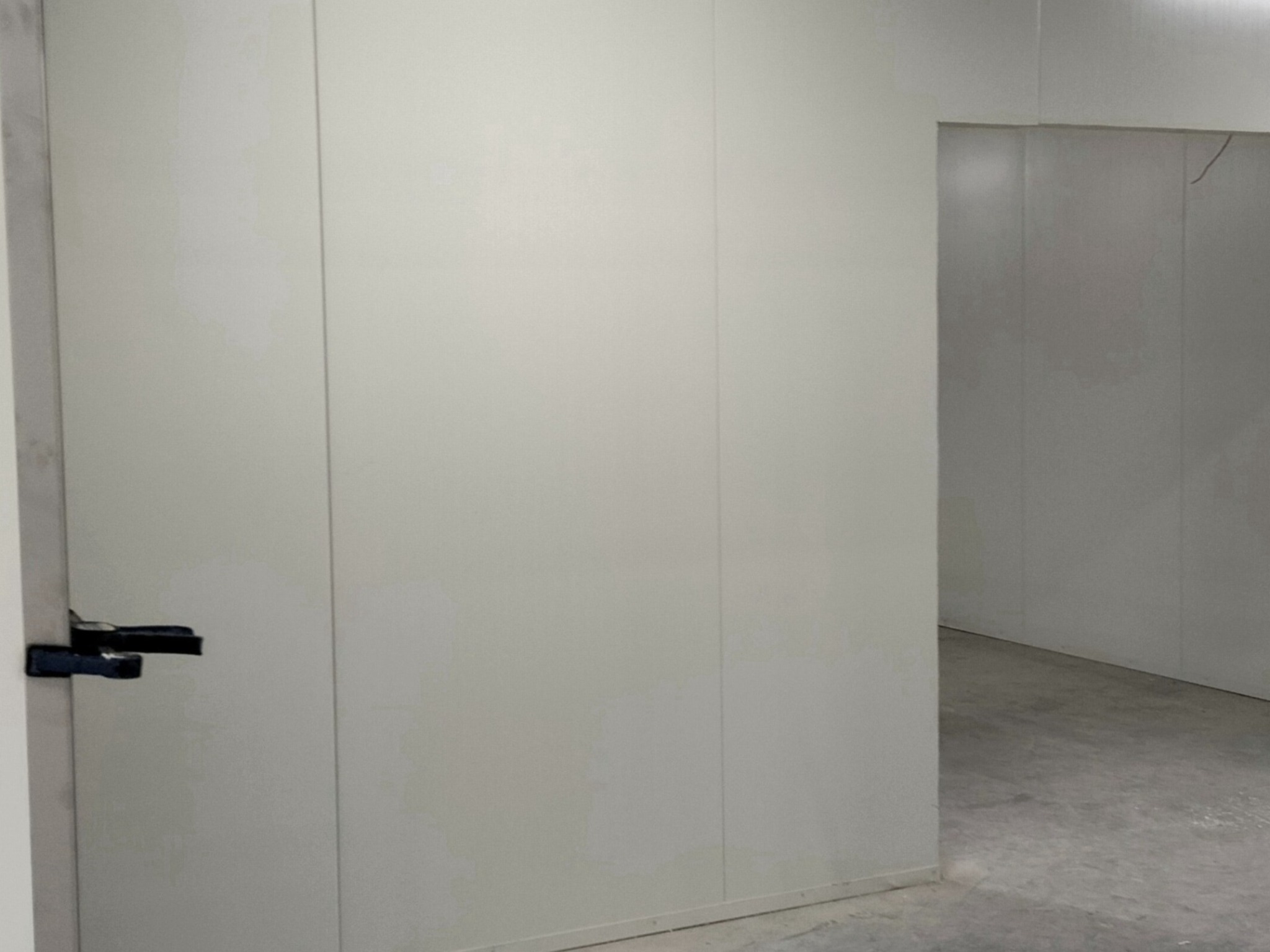 Wnętrze hali przemysłowej z białymi płytami warstwowymi i czarnym uchwytem drzwiowym po lewej stronie kadru, widok na korytarz.