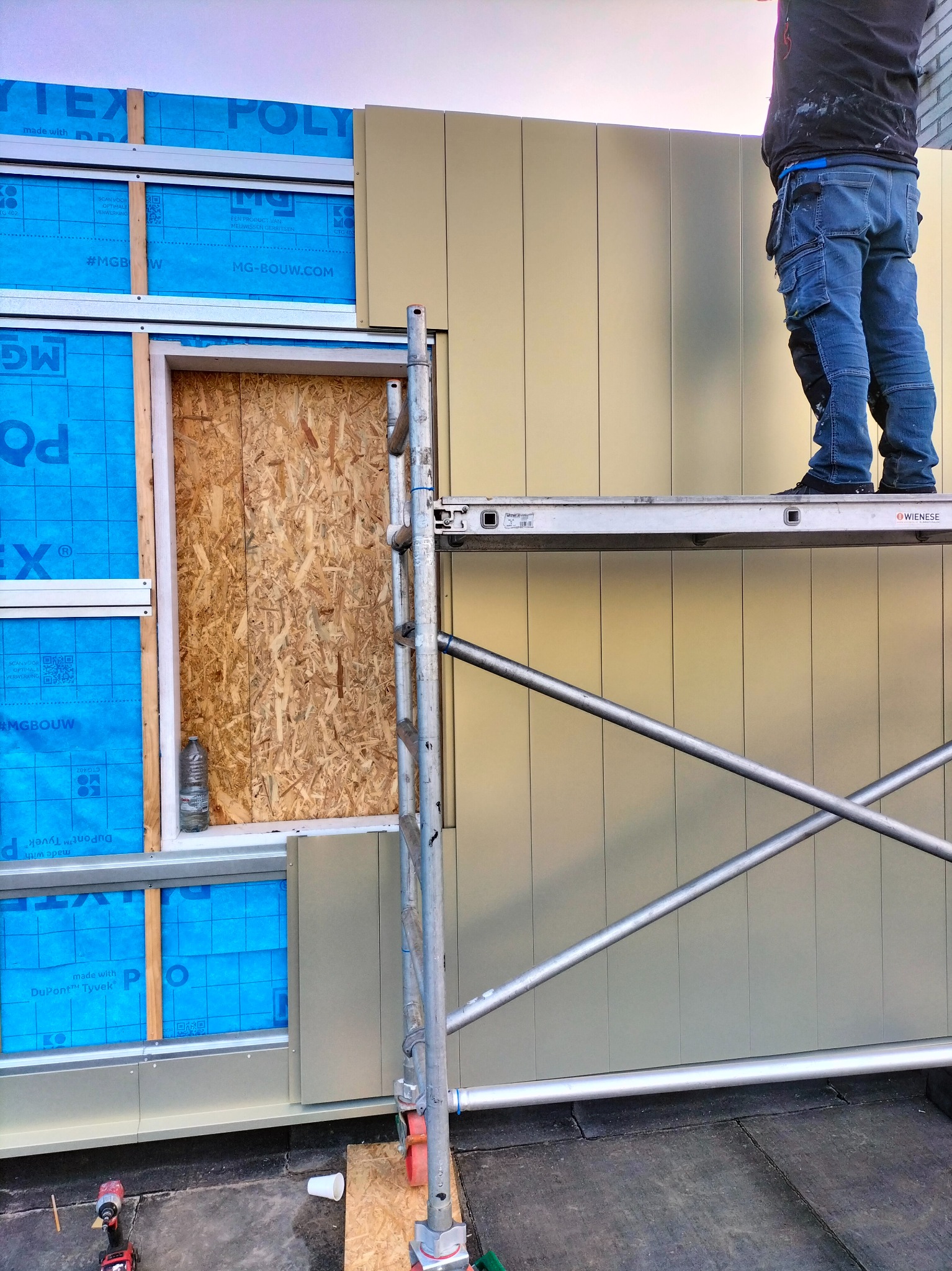 Prace montażowe płyt warstwowych w Szklarach: mężczyzna na rusztowaniu instaluje panele na ścianie budynku, widoczny fragment konstrukcji z izolacją i zabezpieczonym otworem okiennym.