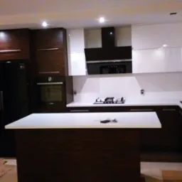 Nowoczesna kuchnia z wyspą, fronty w kolorze białym i ciemnego drewna, czarny sprzęt AGD, widoczne kartony na podłodze sugerujące świeży montaż.