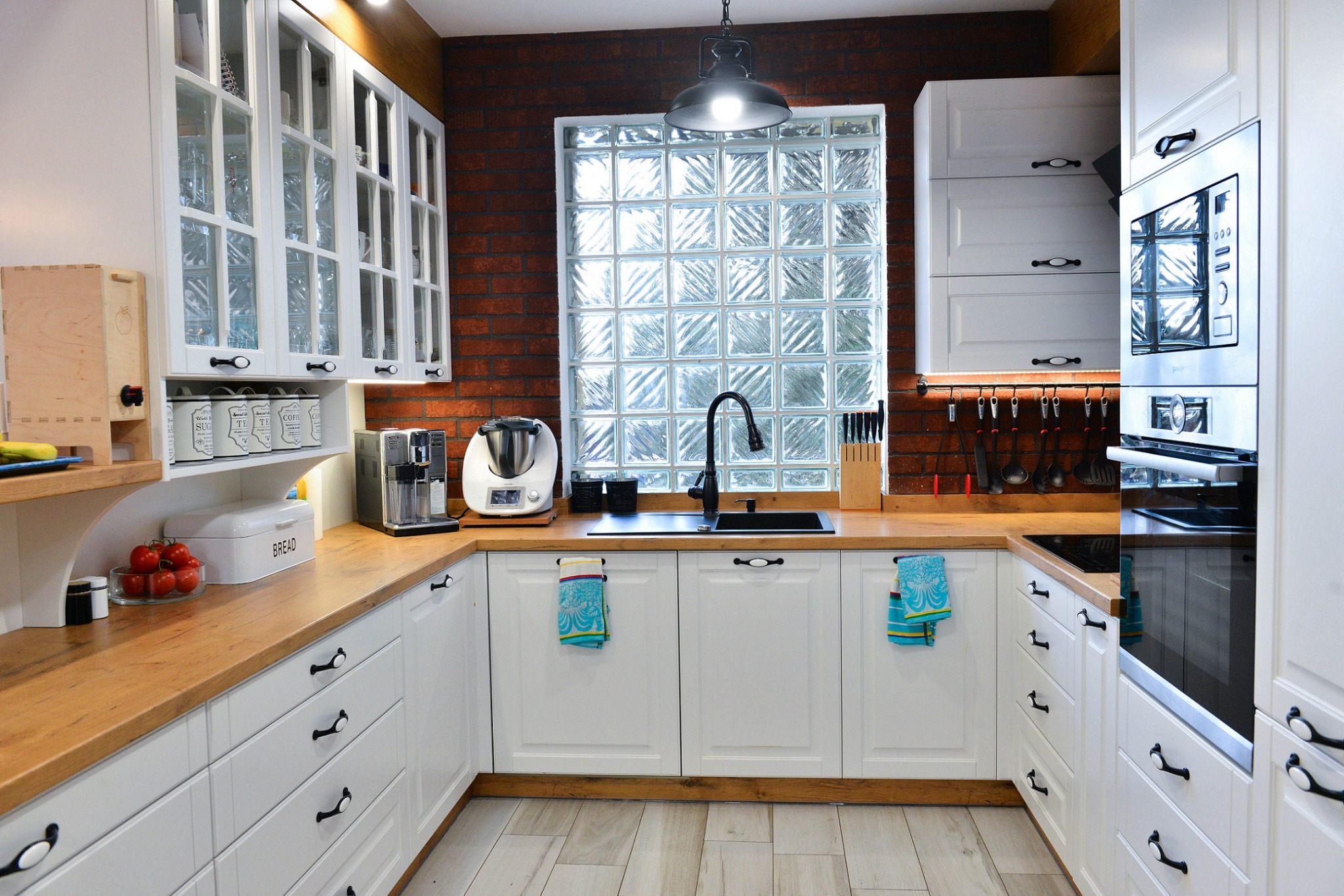 Jasna kuchnia z białymi szafkami i drewnianym blatem, ceglana ściana, okno z luksferów, wisząca lampa i akcesoria kuchenne.