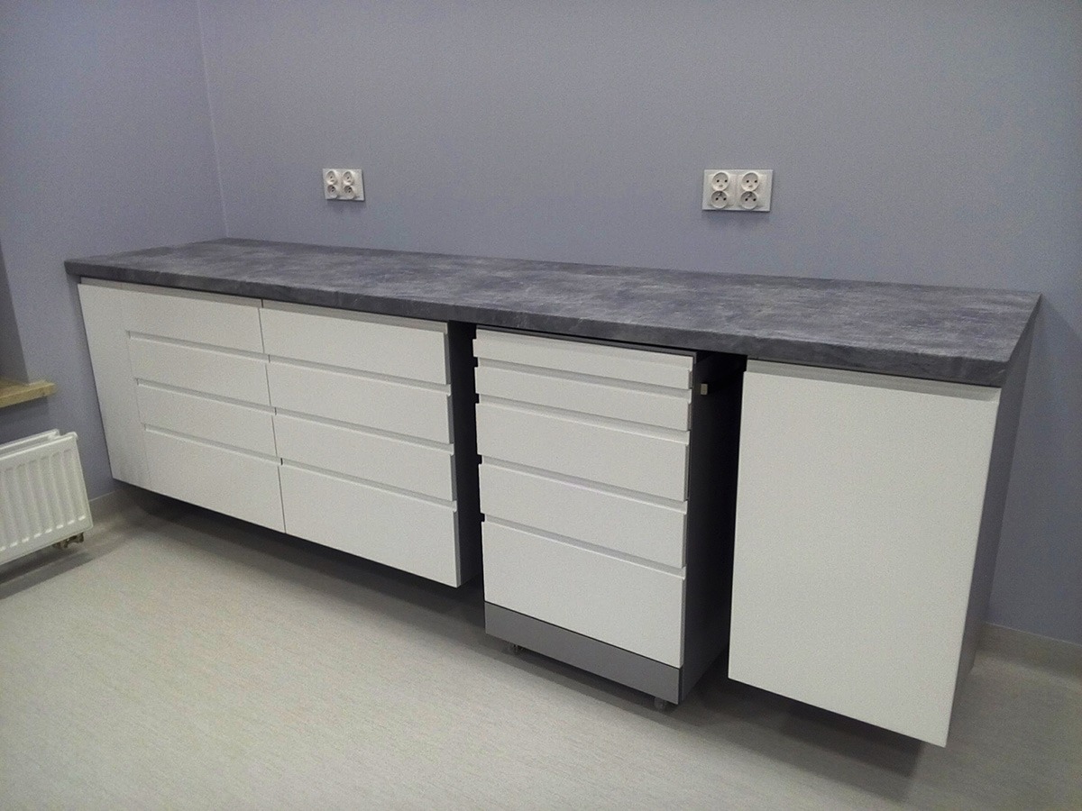 Długa, biała szafka z szufladami i szarym blatem z imitacją kamienia, zamontowana na ścianie w pokoju z szarą ścianą i gniazdkami elektrycznymi.