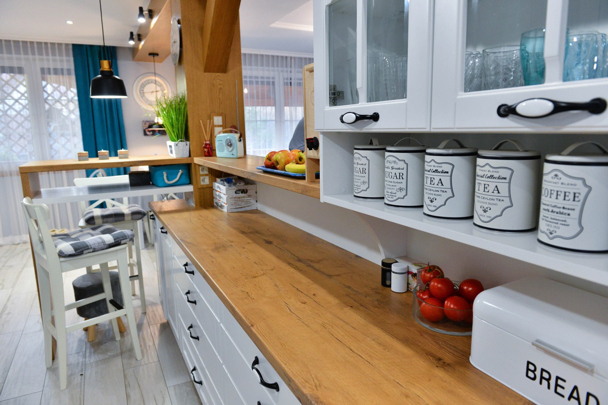 Jasna kuchnia z drewnianym blatem, białymi szafkami z czarnymi uchwytami, dekoracyjnymi puszkami na półce, owocami i pojemnikiem na chleb, widok na jadalnię z turkusowymi akcentami.
