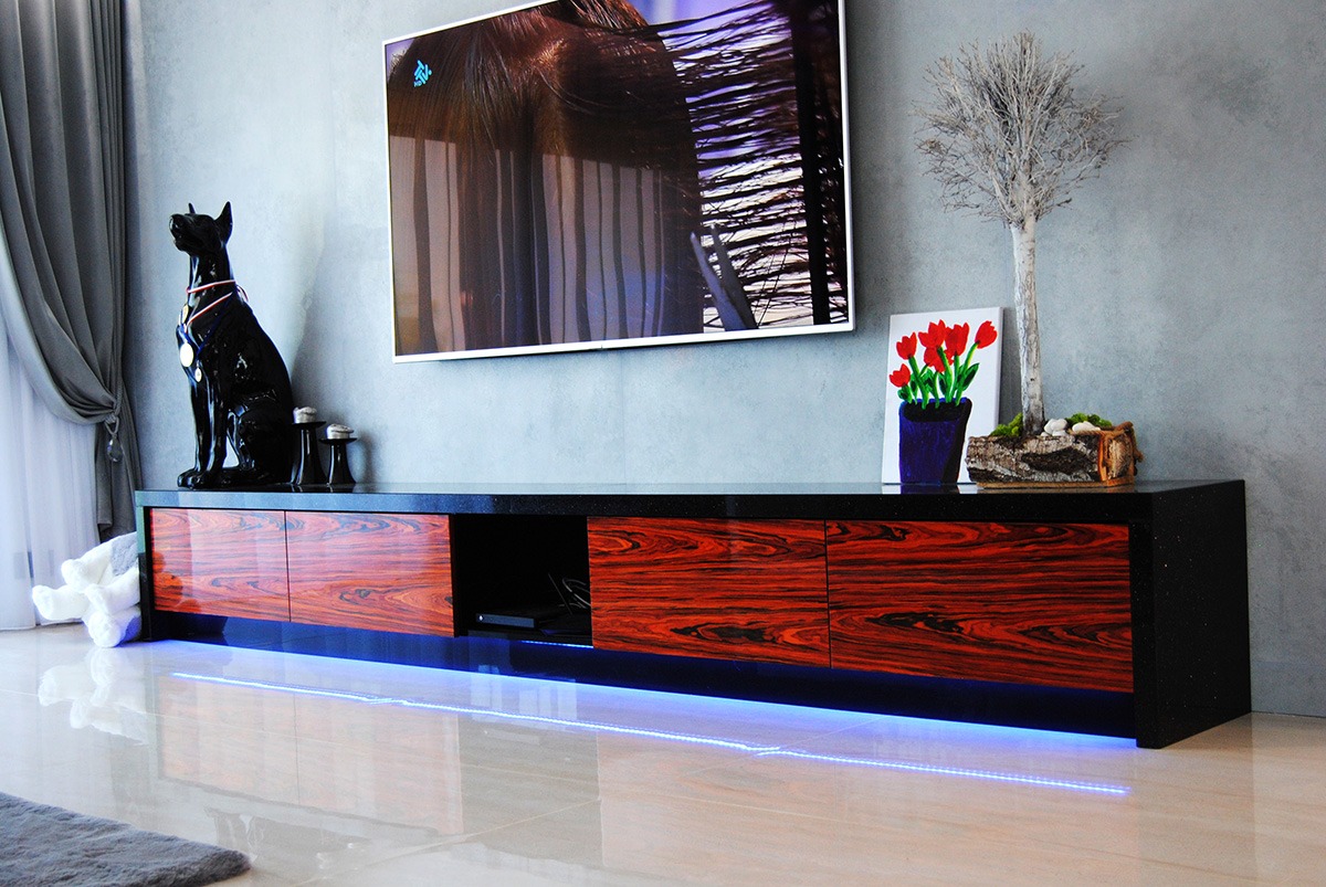 Nowoczesna szafka RTV z fornirem o wzorze palisandru, podświetlona niebieskim światłem LED, z czarnym blatem i dekoracjami: figurka psa, małe wazoniki, obrazek z tulipanami i dekoracyjne drzewko.