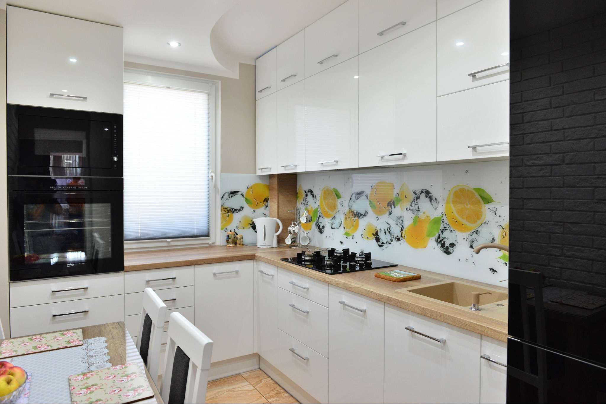 Nowoczesna, jasna kuchnia z białymi, lakierowanymi frontami, drewnianym blatem i szklanym panelem ściennym z motywem cytryn i lodu, widok na płytę grzewczą i zlew.