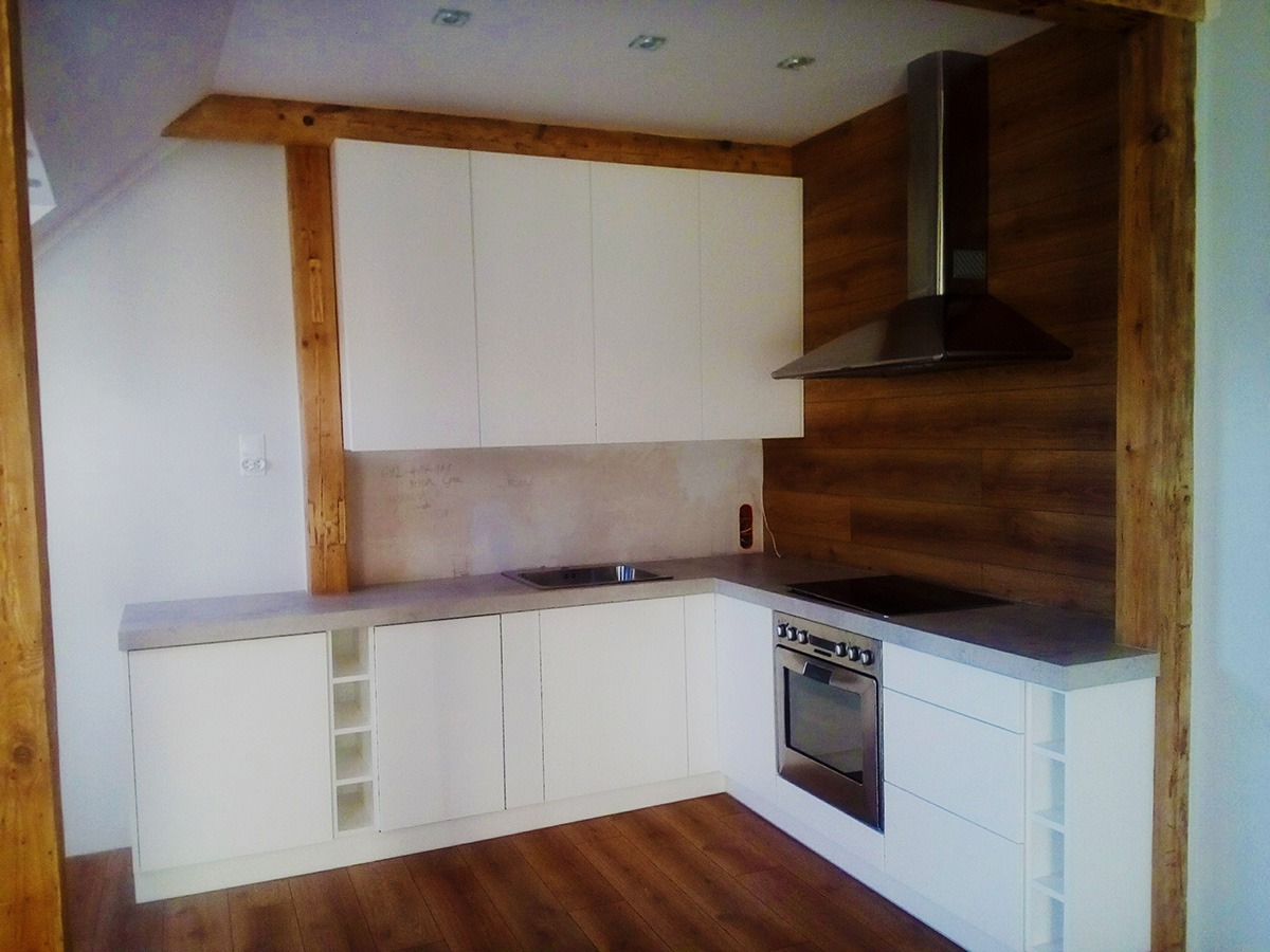 Nowoczesna, biała kuchnia z drewnianymi elementami konstrukcyjnymi, okapem nad płytą indukcyjną i drewnianą podłogą. Na ścianie notatki ołówkiem.