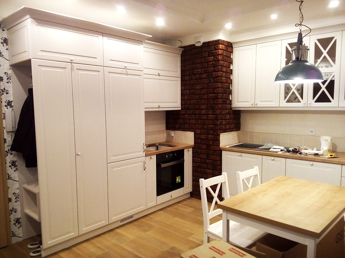 Jasna kuchnia z białymi meblami na wymiar, ceglanym elementem dekoracyjnym i drewnianym stołem z krzesłami w stylu rustykalnym.