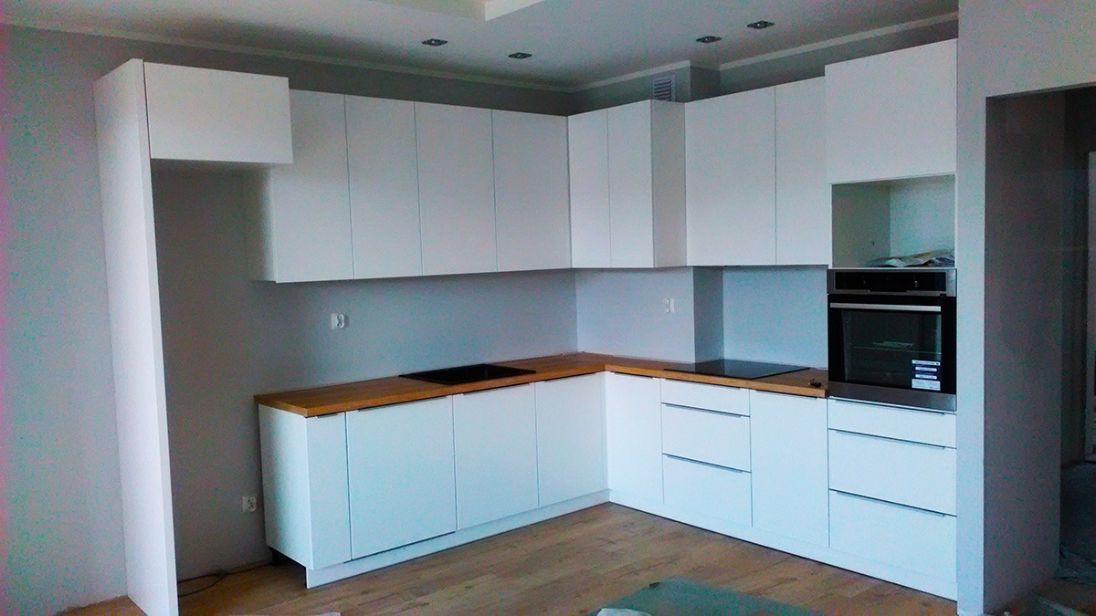 Biała, nowoczesna kuchnia narożna z drewnianym blatem, płyta indukcyjna i piekarnik w zabudowie, widok na fragment pomieszczenia w trakcie wykańczania.