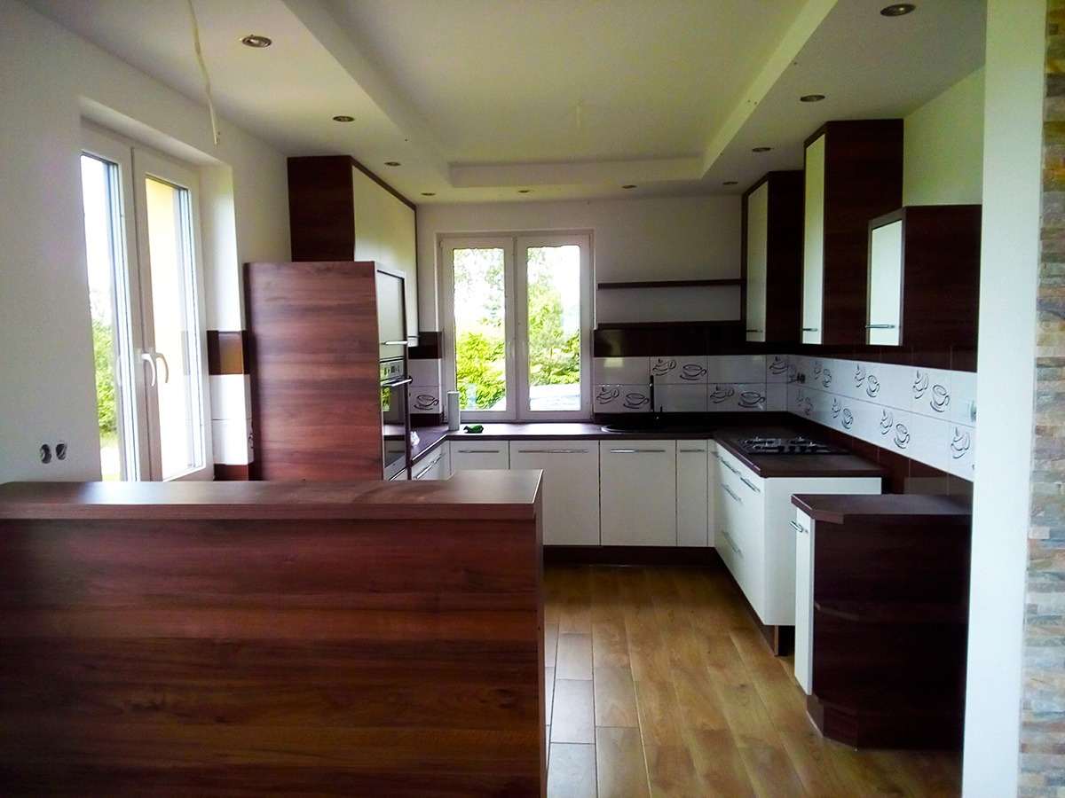 Nowoczesna kuchnia z białymi szafkami dolnymi, ciemnobrązowymi górnymi i blatem, drewniana podłoga, ściana z dekoracyjnymi płytkami z motywem filiżanek, widok na ogród przez okno.