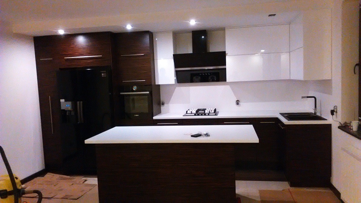 Nowoczesna kuchnia z wyspą, fronty w kolorze białym i ciemnego drewna, czarny sprzęt AGD, widoczne kartony na podłodze sugerujące świeży montaż.