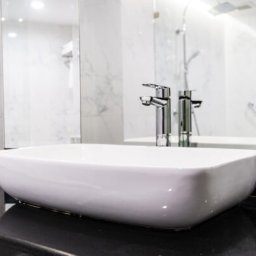 Inspiracje Kamienne - Nowoczesna, prostokątna umywalka nablatowa z baterią chromowaną, ustawiona na czarnym blacie z kamienia, na tle białych marmurowych płytek i lustra z odbiciem kabiny prysznicowej oraz kwiat...