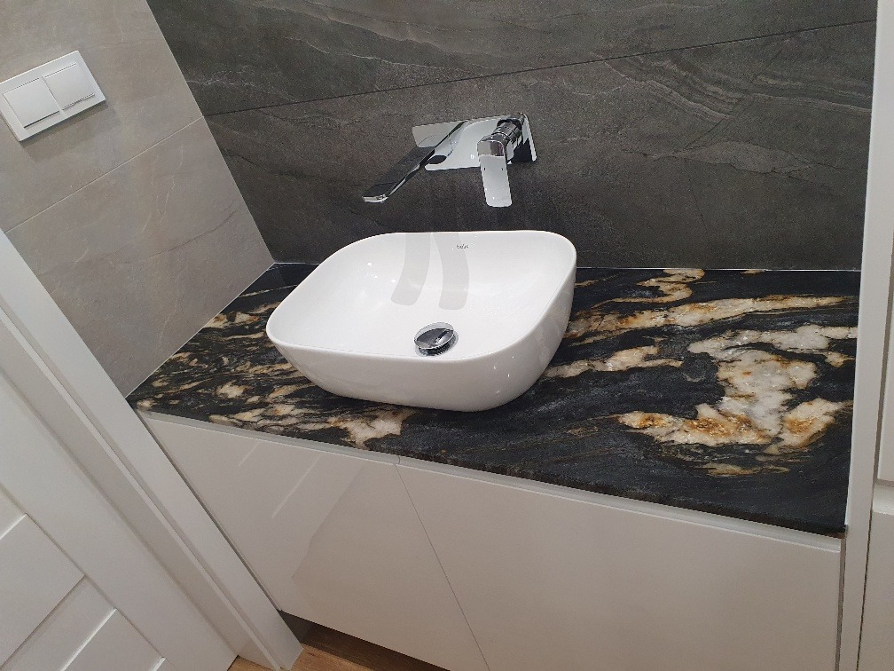 Biała, owalna umywalka nablatowa z chromowaną armaturą osadzona na blacie z czarnego kamienia z wyraźnymi złotymi żyłami, zamontowana na białej szafce w nowoczesnej łazience.