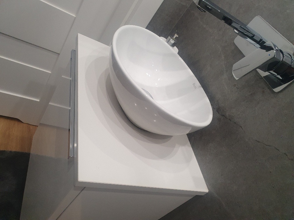 Biała, okrągła umywalka nablatowa marki Rea na białym blacie z szufladami, z chromowaną baterią i szarą posadzką. Widoczny fragment białej ściany z dekoracyjnymi listwami.