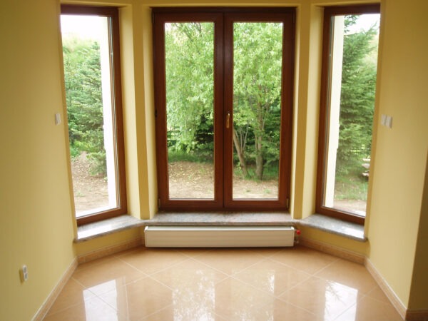 Jasne wnętrze z oknami z drewnianą stolarką i szarymi parapetami, widok na zieleń za oknem, pod oknami grzejnik.