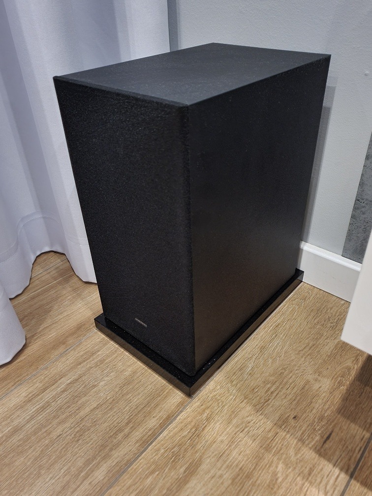 Czarna kolumna głośnikowa na podeście z konglomeratu z drobnymi, błyszczącymi elementami, ustawiona na drewnianej podłodze w rogu pokoju, obok białej zasłony.