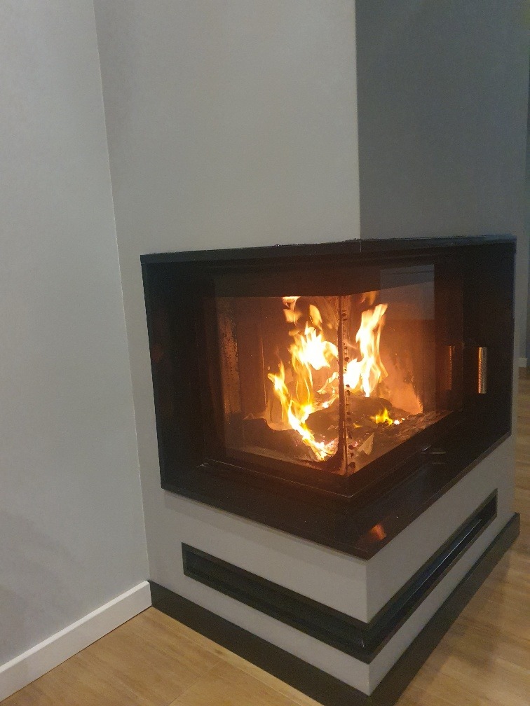 Nowoczesny kominek narożny z obudową w odcieniach szarości i czerni, płomienie tańczące za szybą, widoczne drewno w palenisku, jasna podłoga.