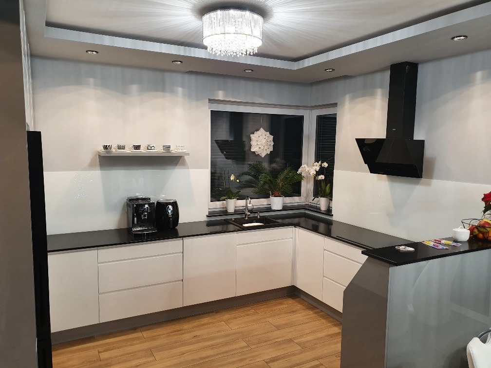 Nowoczesna kuchnia z białymi szafkami, czarnym blatem, drewnianą podłogą i oknem z widokiem na zieleń, oświetlona designerskim żyrandolem i halogenami.