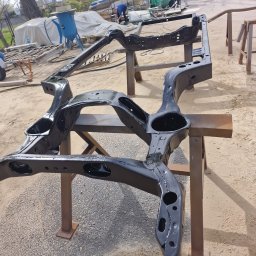 BRUMBUD - Błyszcząca, czarna rama samochodu po renowacji, umieszczona na metalowych kozłach w warsztacie, z widocznym sprzętem do piaskowania i pracującym mężczyzną w tle.