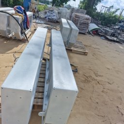 BRUMBUD - Dwa elementy konstrukcyjne pomalowane na szaro, leżące na paletach na piaszczystym podłożu, prawdopodobnie po piaskowaniu i malowaniu proszkowym.