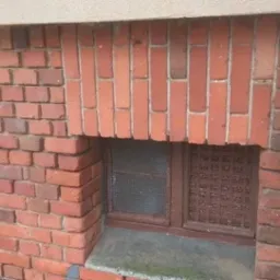Zamknięte okienko piwniczne z kratką, wmurowane w ceglany mur budynku, z widoczną rurą spustową po lewej stronie i fragmentem rusztowania po prawej.